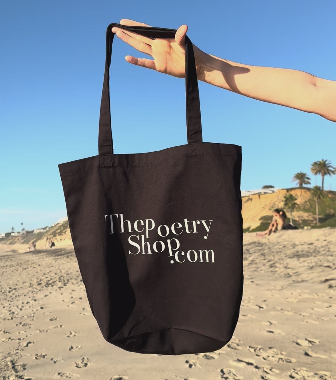 video of poetry shop tote bag held in the air by a woman's hand and gently swaying in the breeze on a southern california beach